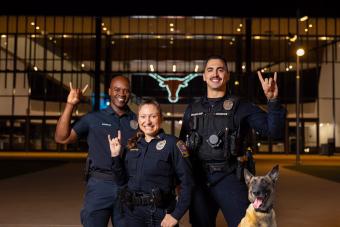Three UTPD officers and their canine pose with horns up outside of Moody Center